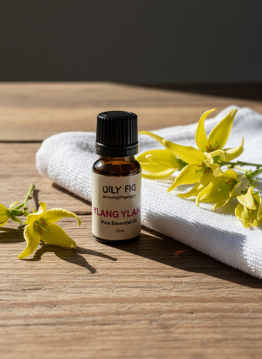 Oily Fig Aromatherapy Ylang Ylang essential oil bottle on a wooden surface with yellow flowers and a white towel.